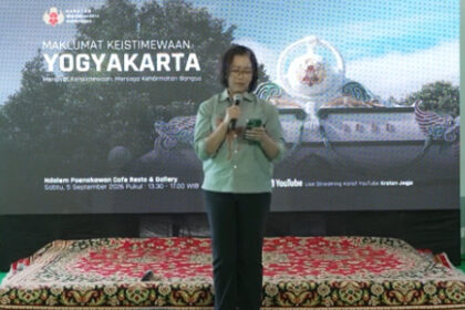 Female presenter on a stage, holding a microphone, in front of a banner that reads 'Maklumat Keistimewaan Yogyakarta' with decorative backdrop and a patterned carpet.