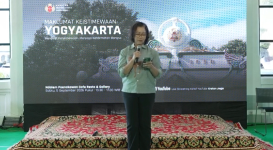 Female presenter on a stage, holding a microphone, in front of a banner that reads 'Maklumat Keistimewaan Yogyakarta' with decorative backdrop and a patterned carpet.