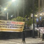 Banner in Indonesian protesting, reading: 'WARGA JOGJA MENOLAK KERAS DEMONSTRASI YANG MENGGANGU AKTIVITAS & KETERTIBAN UMUM' displayed along a black fence at night with street lamps and decorative posts.