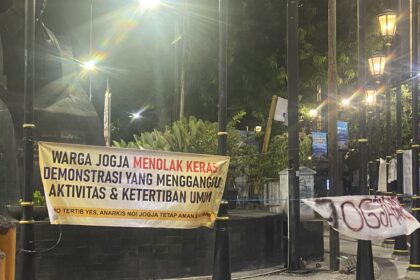 Banner in Indonesian protesting, reading: 'WARGA JOGJA MENOLAK KERAS DEMONSTRASI YANG MENGGANGU AKTIVITAS & KETERTIBAN UMUM' displayed along a black fence at night with street lamps and decorative posts.