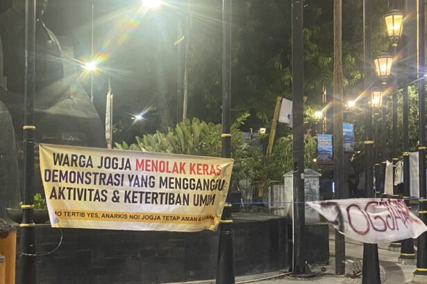 Banner in Indonesian protesting, reading: 'WARGA JOGJA MENOLAK KERAS DEMONSTRASI YANG MENGGANGU AKTIVITAS & KETERTIBAN UMUM' displayed along a black fence at night with street lamps and decorative posts.