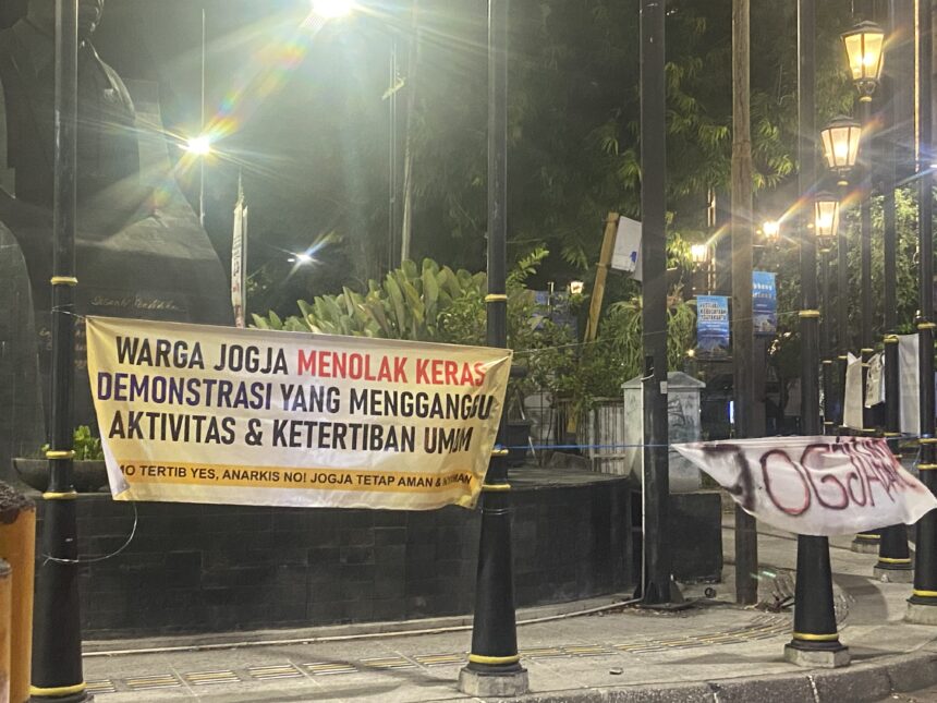 Banner in Indonesian protesting, reading: 'WARGA JOGJA MENOLAK KERAS DEMONSTRASI YANG MENGGANGU AKTIVITAS & KETERTIBAN UMUM' displayed along a black fence at night with street lamps and decorative posts.