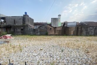 Dry, stubbled field in foreground with weathered brick buildings and a decorative tall clock-tower on the horizon under a blue sky.