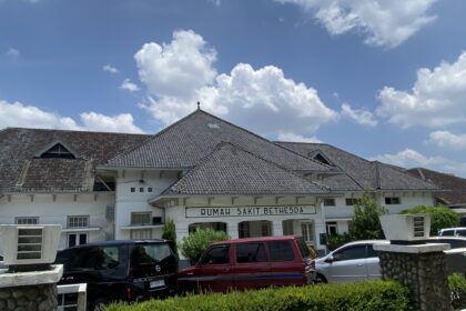 Front view of Rumah Sakit Bethesda hospital with white facade and tiled roof under a blue sky.