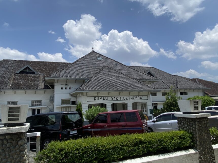 Front view of Rumah Sakit Bethesda hospital with white facade and tiled roof under a blue sky.