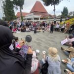 Large group of people gathered outdoors for a protest or rally, some seated and some standing, holding signs and recording with phones.