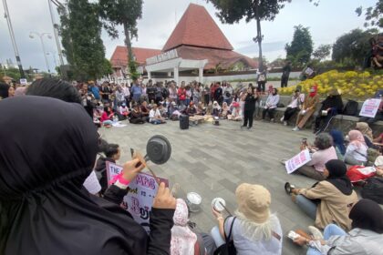 Large group of people gathered outdoors for a protest or rally, some seated and some standing, holding signs and recording with phones.