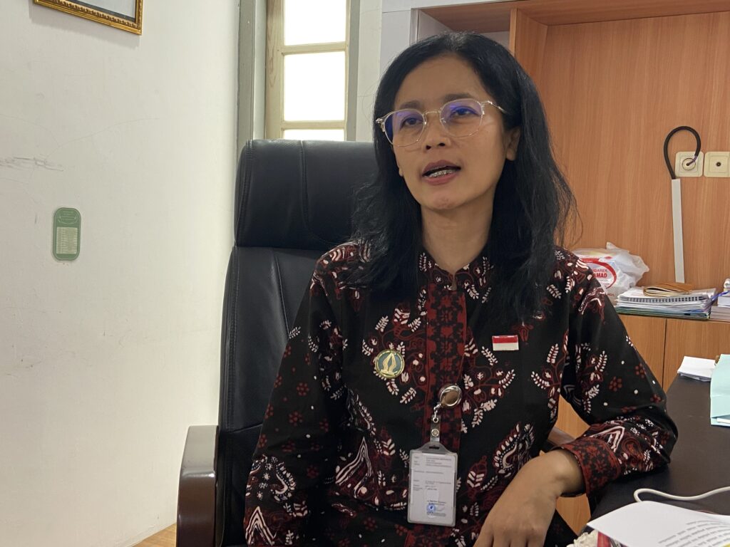 IMG_3438 1 - Mabur.co Professional woman seated at a desk in an office, wearing glasses and a patterned red‑black top with an ID badge visible on a lanyard.