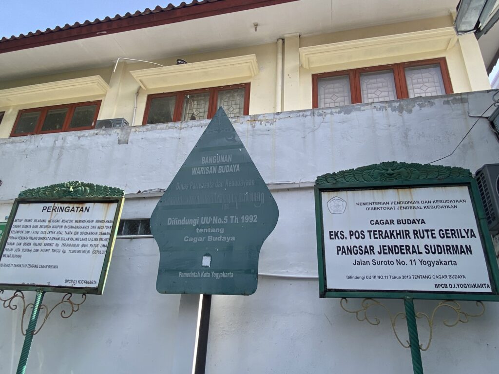 IMG_3443 1 - Mabur.co Signboards with Indonesian government notices about cultural heritage mounted on a pale wall in front of a building. Framed red windows above and decorative green metalwork visible.