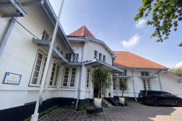White two-story building with blue-gray trim and red-tiled roofs in a sunny courtyard, potted plants at the entrance, and a parked black SUV nearby.