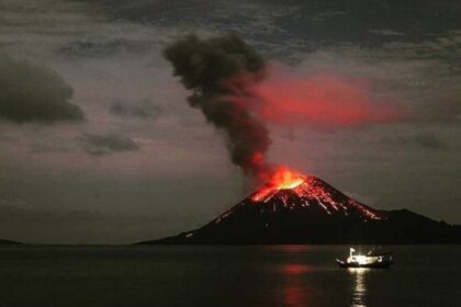 Volcanic eruption at night with lava spewing from the crater and dark smoke rising, a small boat on calm water in the foreground.