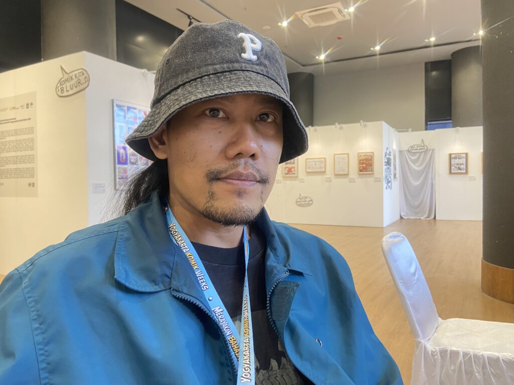 Self-portrait of a man in a blue jacket wearing a gray bucket hat with a lanyard around his neck, in an exhibition space with white walls and displays behind him.