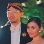 Man in a tuxedo with a cream bow tie and glasses standing with a woman in a pink dress at night.