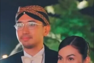Man in a tuxedo with a cream bow tie and glasses standing with a woman in a pink dress at night.