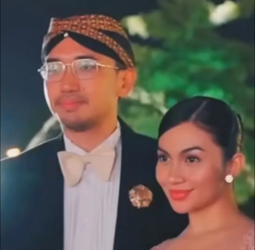 Man in a tuxedo with a cream bow tie and glasses standing with a woman in a pink dress at night.