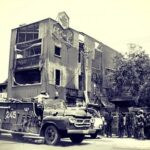 War-torn street with a damaged, multi-story building and an armored vehicle parked in front amid onlookers.