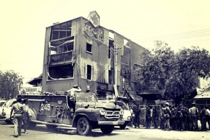 War-torn street with a damaged, multi-story building and an armored vehicle parked in front amid onlookers.
