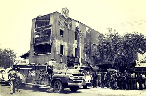 War-torn street with a damaged, multi-story building and an armored vehicle parked in front amid onlookers.