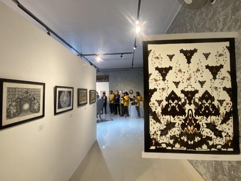 Art gallery interior with framed works along the left wall and a large black-and-white patterned textile piece on the right; group of visitors in yellow shirts in the background.