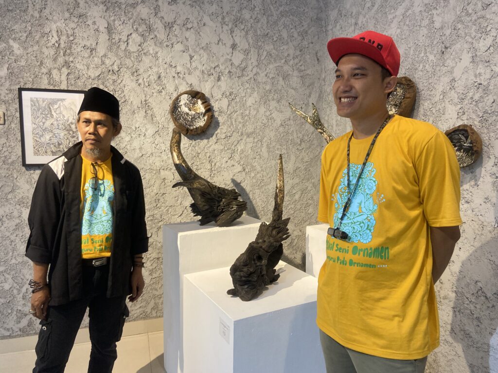 Two men in yellow shirts pose beside intricate metal sculptures on white pedestals in a gallery environment.