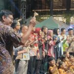 Large group of people in colorful batik shirts on a stage, raising small goblets in a toast at an outdoor event.