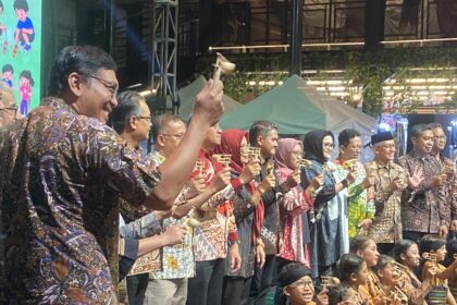 Large group of people in colorful batik shirts on a stage, raising small goblets in a toast at an outdoor event.
