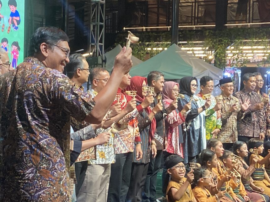 Large group of people in colorful batik shirts on a stage, raising small goblets in a toast at an outdoor event.