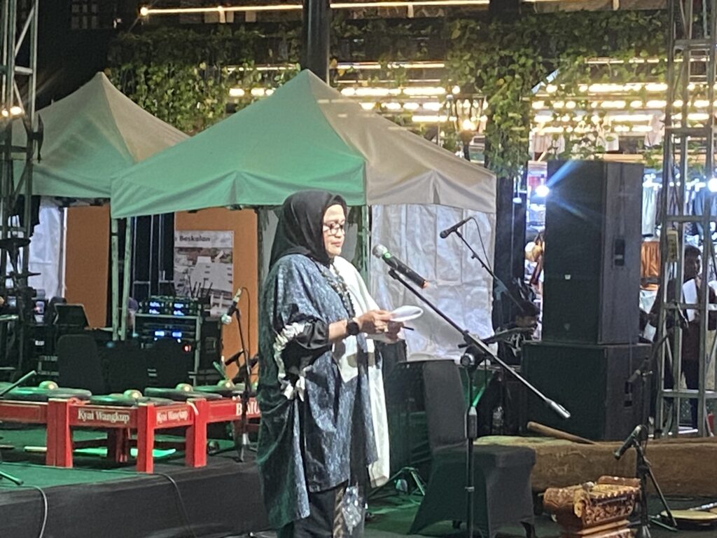 IMG_3704 - Mabur.co Woman in a hijab speaks into a microphone on a dimly lit outdoor stage, with green tents behind her and equipment around her.