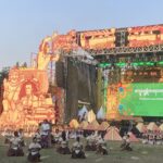 Open-air festival stage with large orange sculpted props and a corps of dancers performing on the grass in white tops and brown skirts.