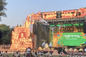 Open-air festival stage with large orange sculpted props and a corps of dancers performing on the grass in white tops and brown skirts.