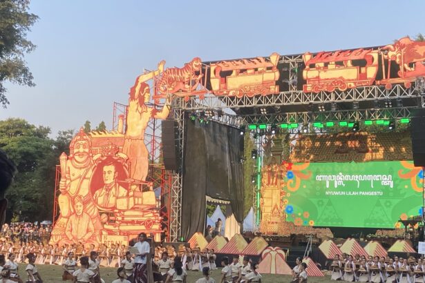 Open-air festival stage with large orange sculpted props and a corps of dancers performing on the grass in white tops and brown skirts.