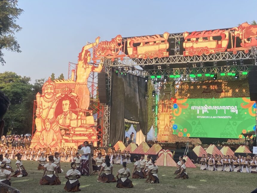 Open-air festival stage with large orange sculpted props and a corps of dancers performing on the grass in white tops and brown skirts.
