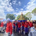 A street procession with performers in red and purple skirts carrying a tall flower-adorned ceremonial frame.