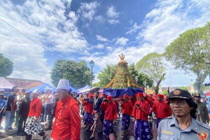 A street procession with performers in red and purple skirts carrying a tall flower-adorned ceremonial frame.