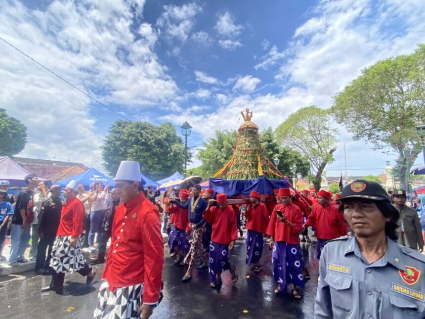 A street procession with performers in red and purple skirts carrying a tall flower-adorned ceremonial frame.