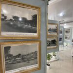 Ornate gold-framed black-and-white photos on a gray display wall in a museum, with a visitor in hijab standing nearby.