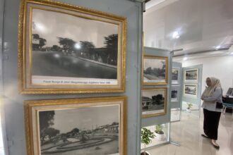 Ornate gold-framed black-and-white photos on a gray display wall in a museum, with a visitor in hijab standing nearby.
