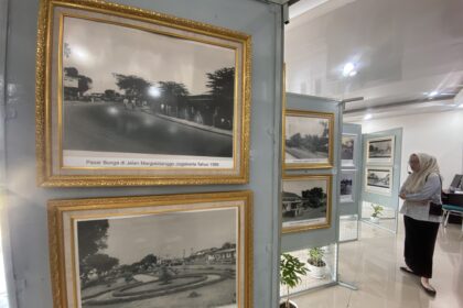 Ornate gold-framed black-and-white photos on a gray display wall in a museum, with a visitor in hijab standing nearby.