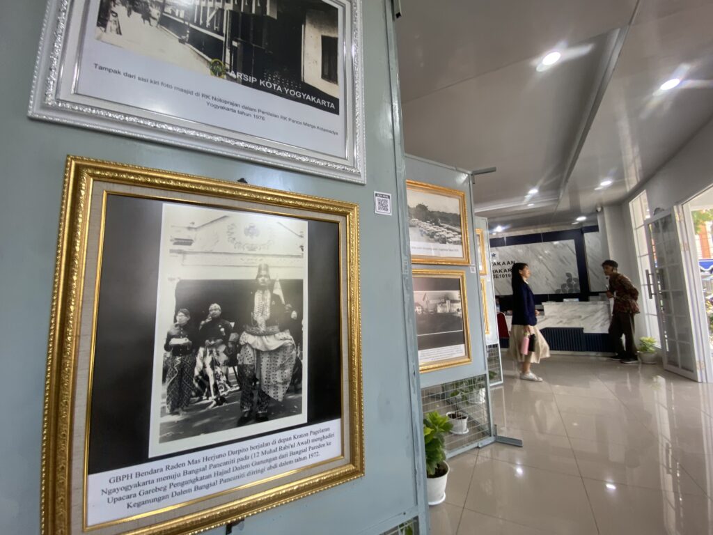 Museum exhibit wall with a large silver-framed black-and-white photo and Indonesian caption beneath, gold-framed photos nearby.