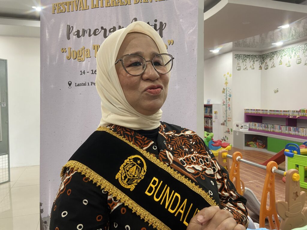 Woman wearing a beige hijab and round glasses, with a black and orange patterned outfit and a gold-embellished sash at a literacy festival backdrop.