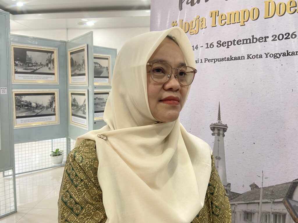Woman wearing a beige hijab and glasses at an art exhibit, with framed photos on blue display panels in the background.
