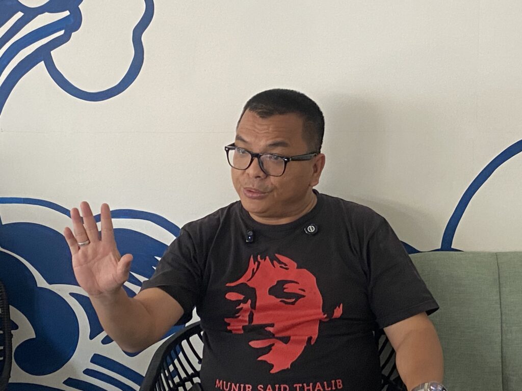 Man in a black shirt with a red dragon graphic waving his left hand, seated indoors against a white wall with blue abstract mural.