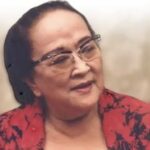 Portrait of an older woman wearing glasses and a red patterned blouse, looking to the side with a gentle smile.