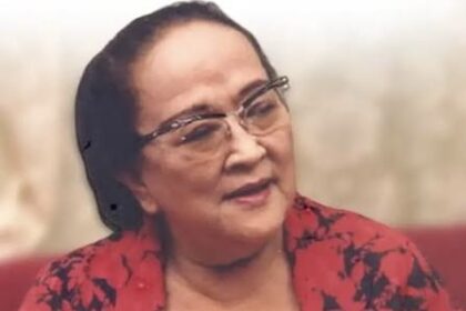 Portrait of an older woman wearing glasses and a red patterned blouse, looking to the side with a gentle smile.