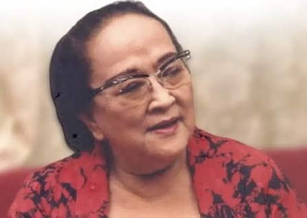 Portrait of an older woman wearing glasses and a red patterned blouse, looking to the side with a gentle smile.