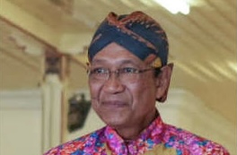 Indonesian man wearing a bright batik shirt and traditional head wrap, smiling indoors at an event.