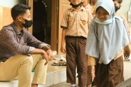 Man wearing a face mask sits on steps and talks with students in uniform outside a building.