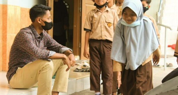 Man wearing a face mask sits on steps and talks with students in uniform outside a building.