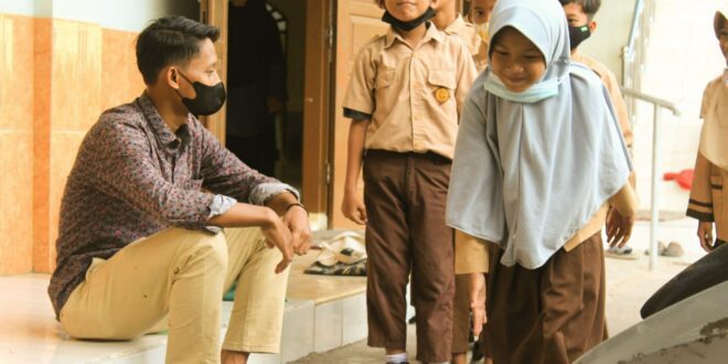 Man wearing a face mask sits on steps and talks with students in uniform outside a building.