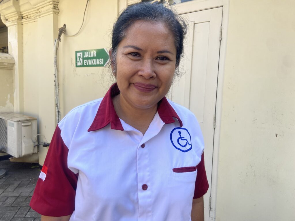 Smiling woman in a white and red uniform standing outside, with a green evacuation sign in the background.